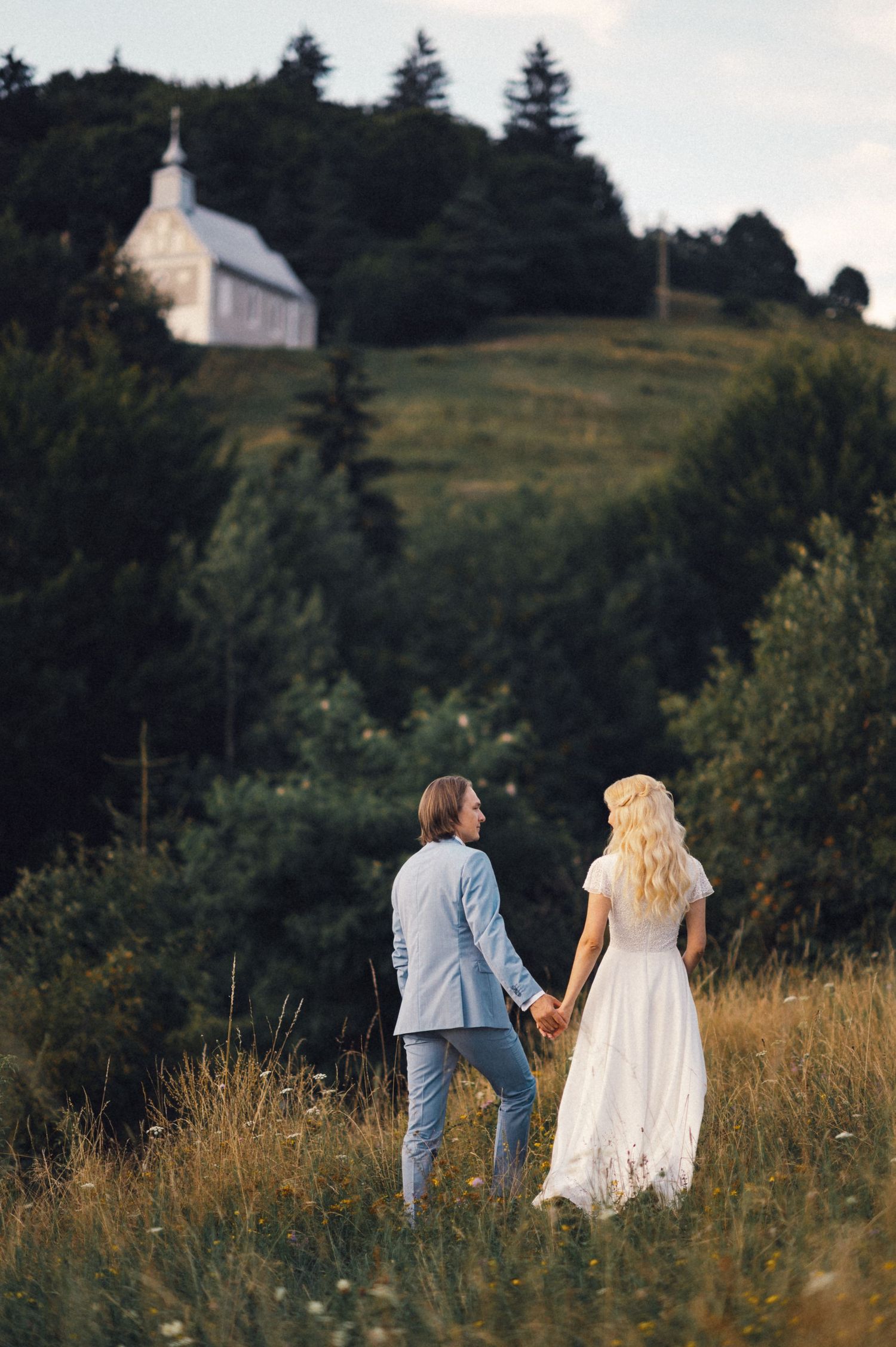 Couple holding hands while walking towards a hilltop chapel surrounded by pine trees.