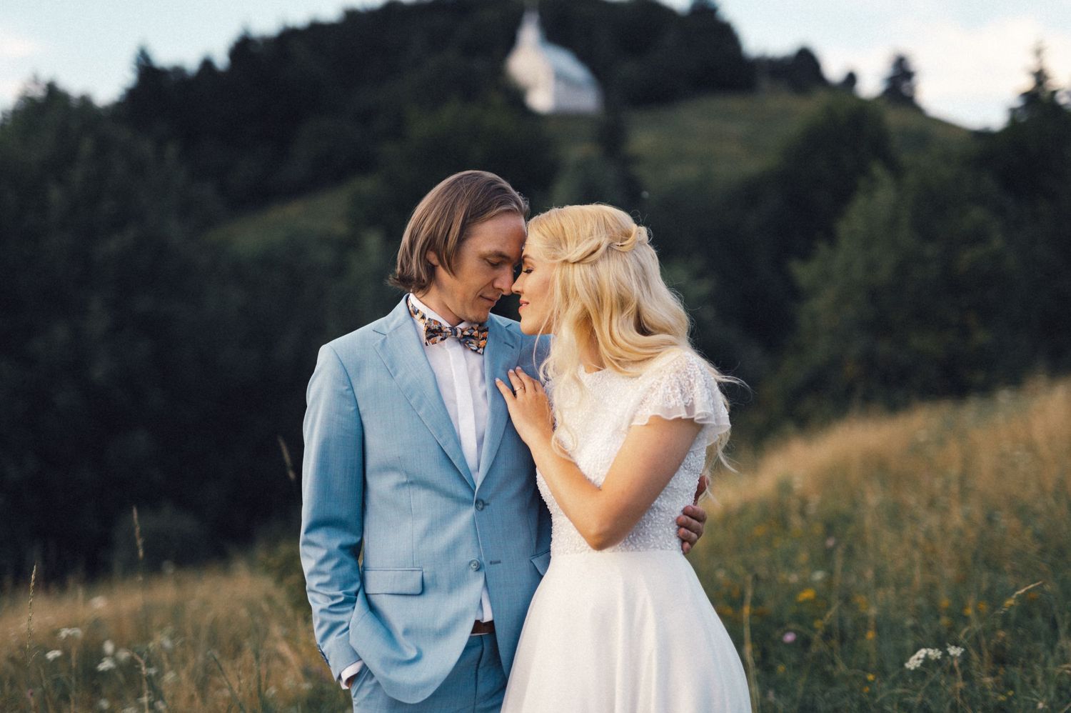 A romantic outdoor wedding photo of a couple in a light blue suit and white dress against a forested hillside.