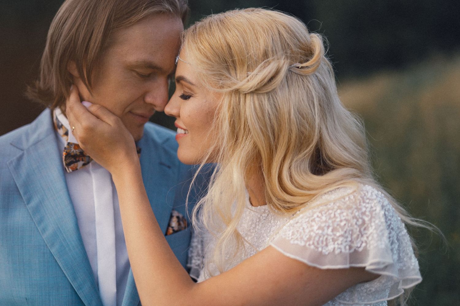 Intimate wedding portrait showing tender moment between couple in blue suit and lace dress outdoors.