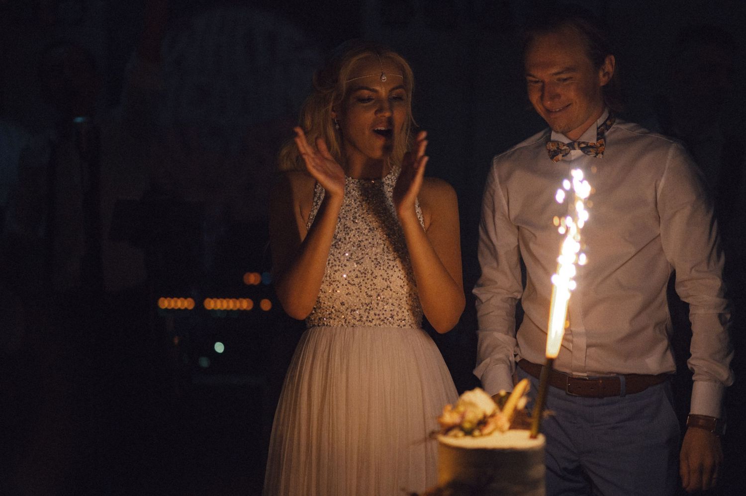 Wedding couple reacting to sparkler-lit cake during evening reception celebration.