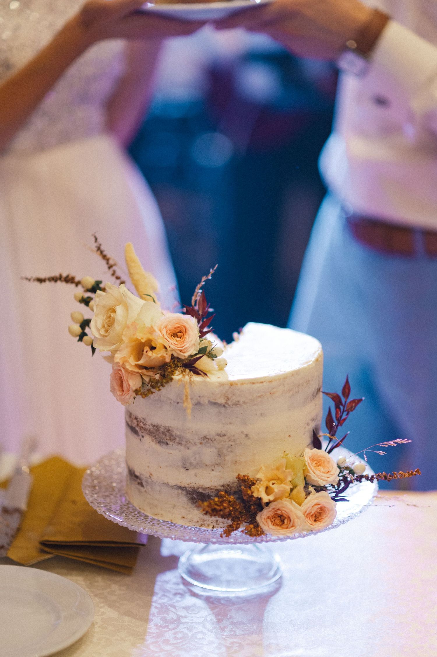 A semi-naked wedding cake decorated with peach roses and delicate foliage on a glass cake stand.