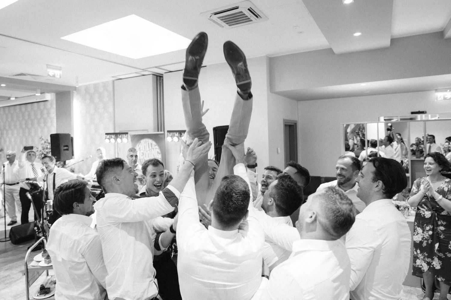 Wedding guests lift someone up during a celebratory moment at an indoor reception.