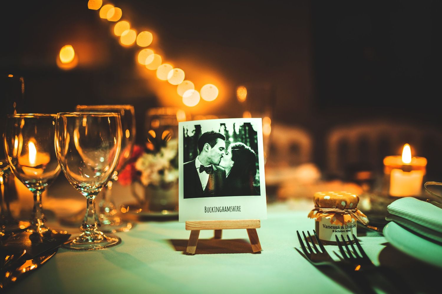 Wedding reception table setting with wine glasses and polaroid photo display illuminated by string lights.
