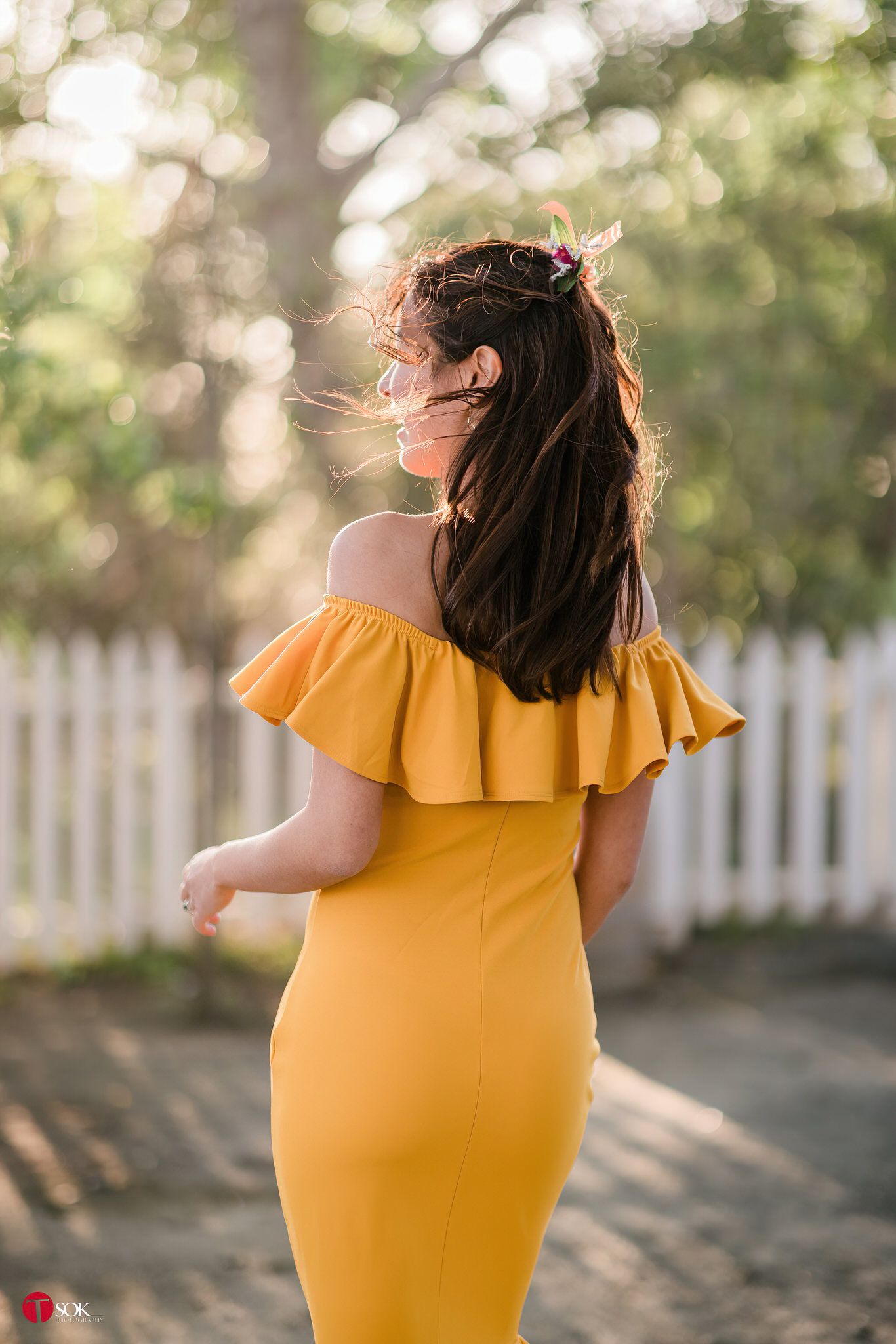 Back view of a woman in a yellow ruffle dress standing against a white fence in sunset lighting.