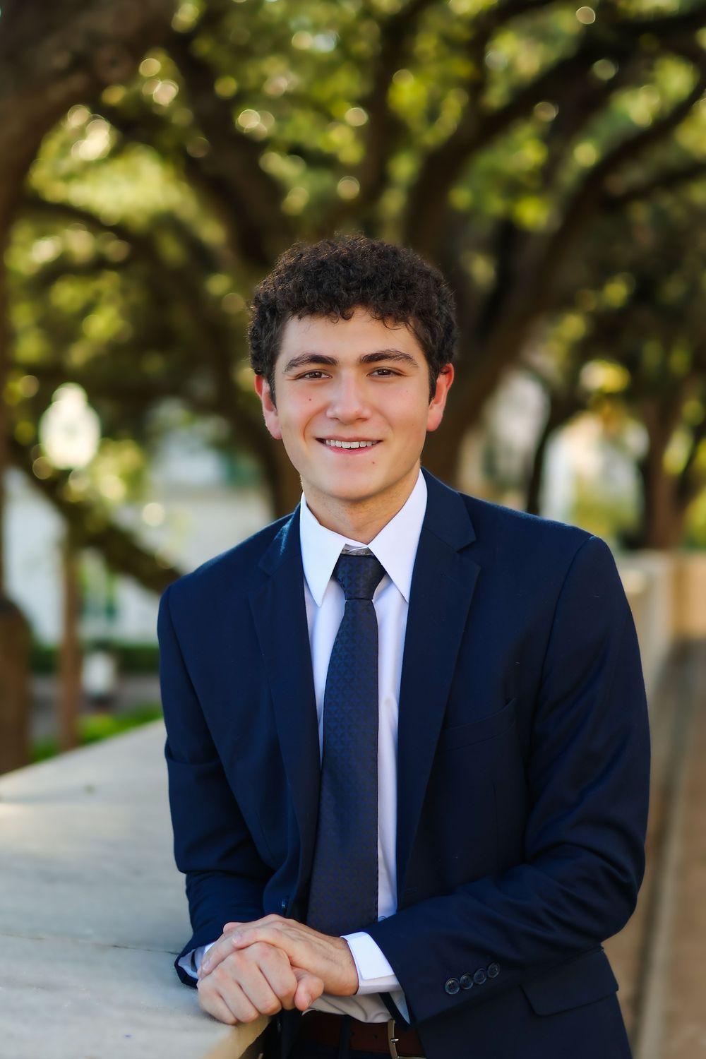 Professional headshot of young professional in navy suit against tree-lined backdrop.