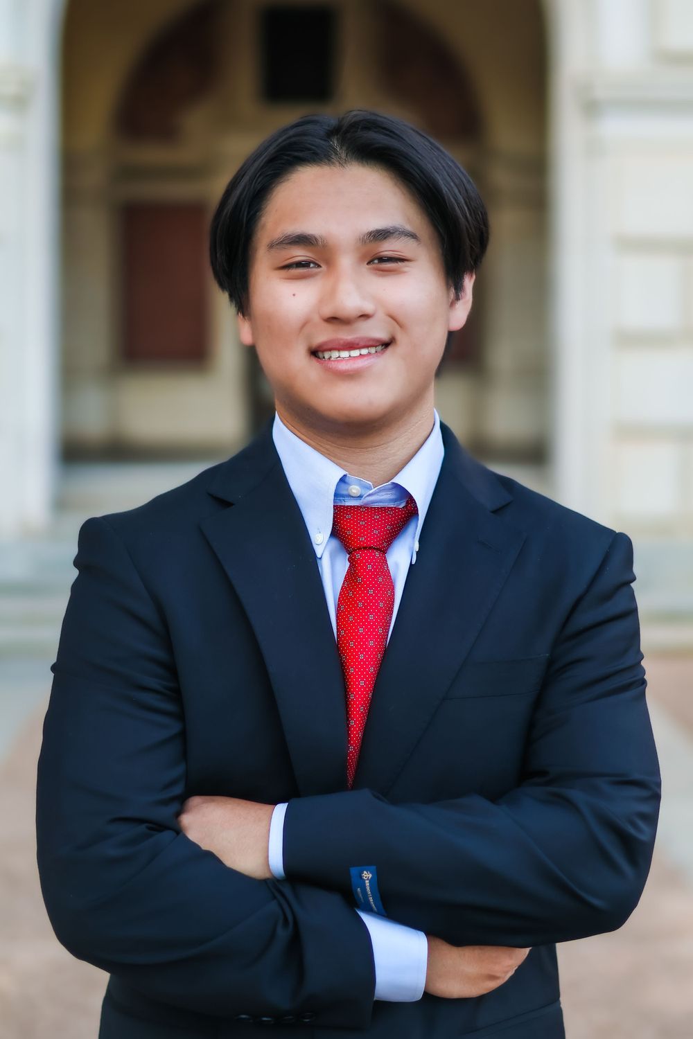 Professional portrait in dark suit with red tie and crossed arms against architectural background.