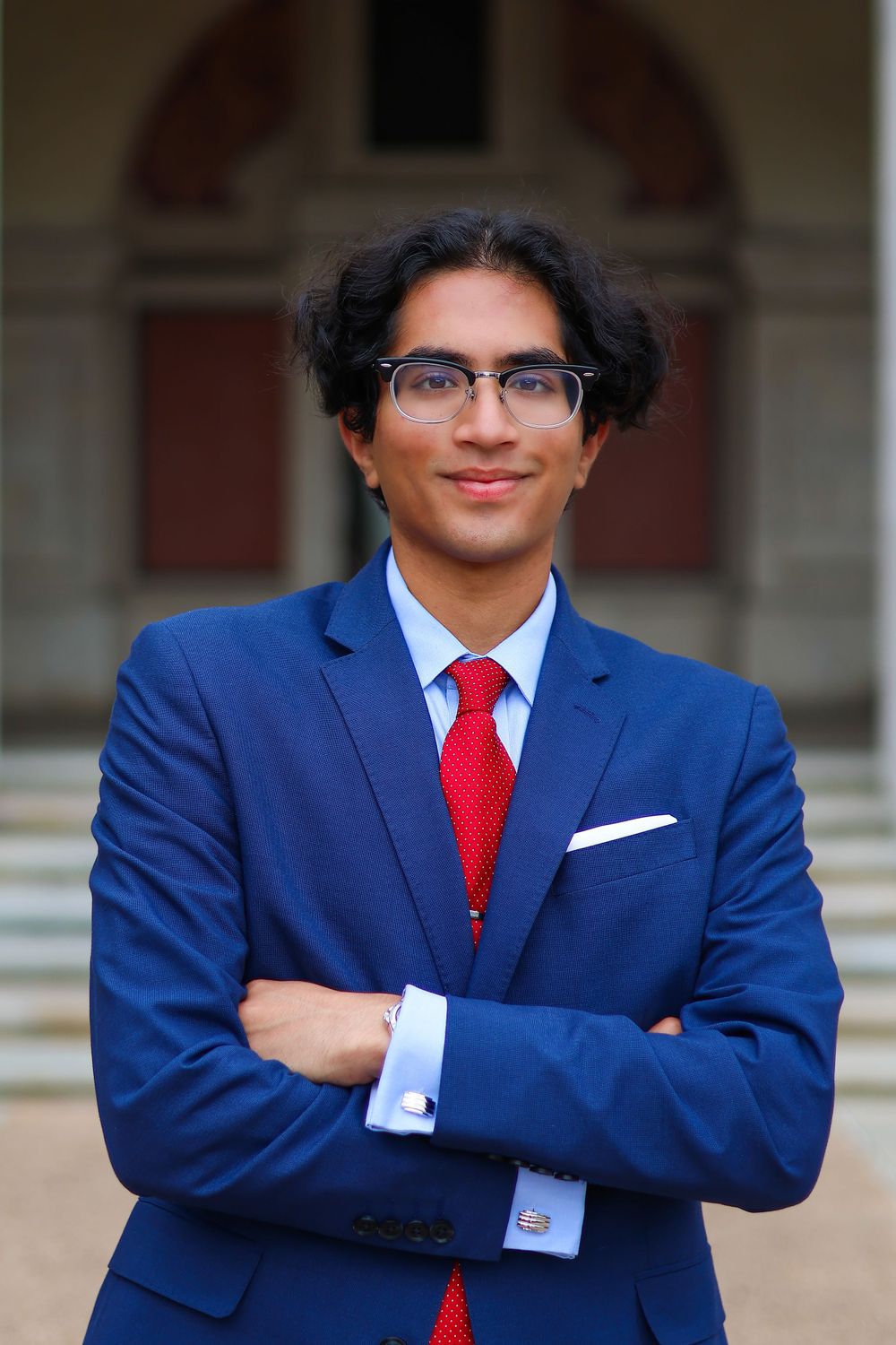 Professional portrait in bright blue suit with red tie and glasses against classic architecture.