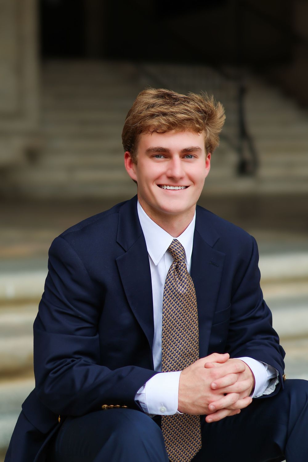 Professional portrait in navy blazer seated on steps with hands clasped.