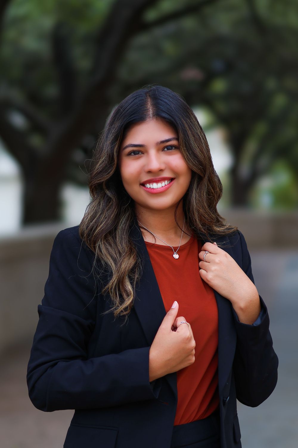 Professional portrait in black blazer and rust-colored top against outdoor backdrop.