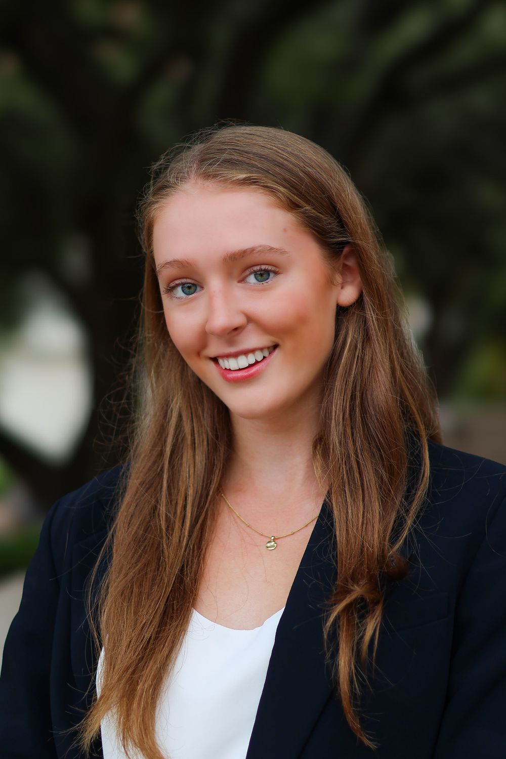 Professional portrait in navy blazer and white top with delicate necklace against natural background.