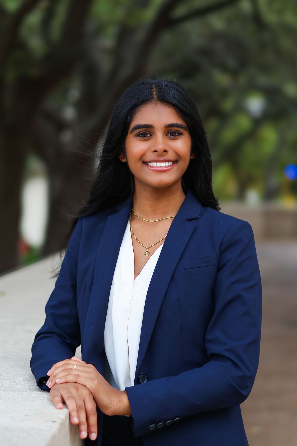 Professional portrait in navy blazer and white blouse with layered necklaces against tree background.