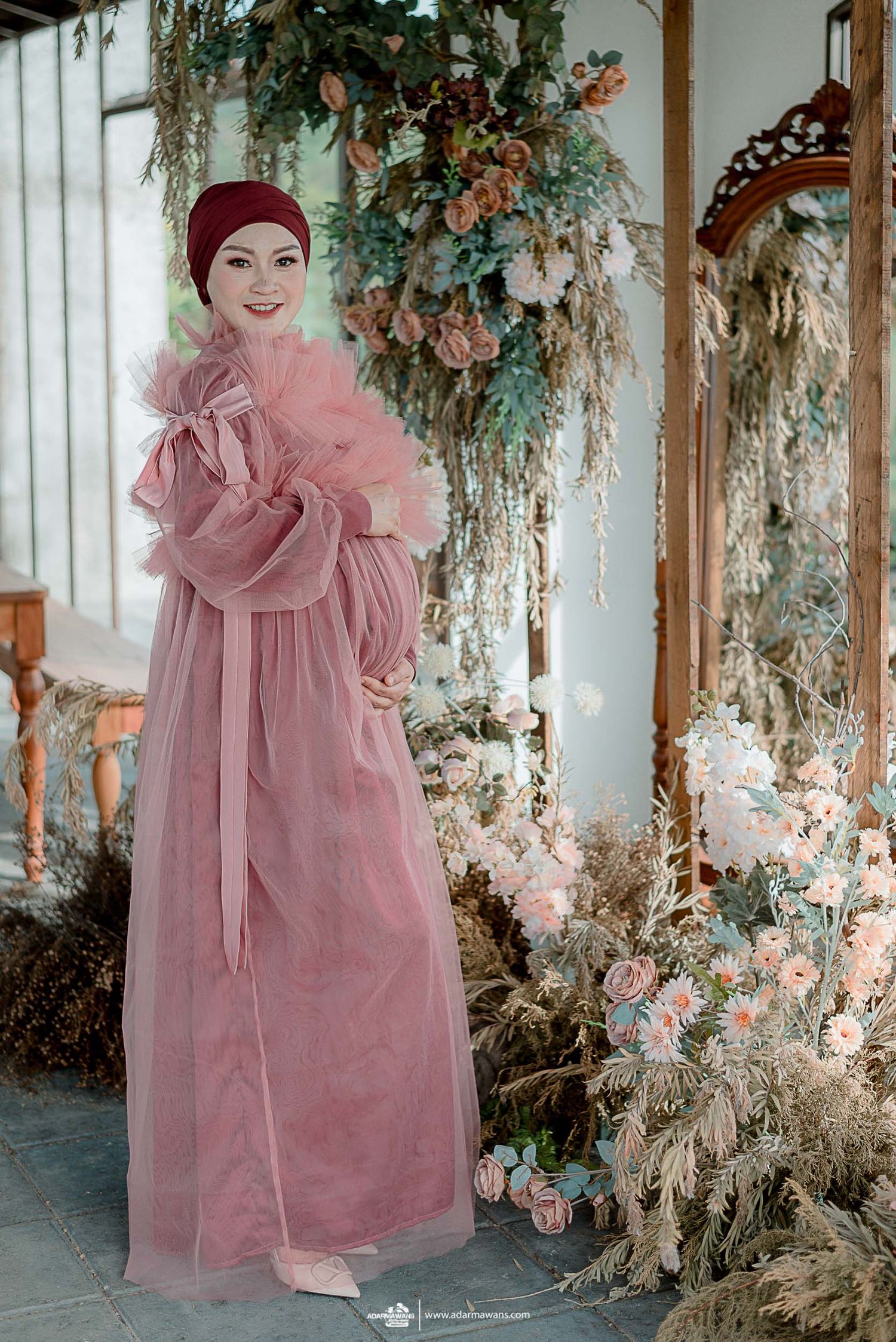 Person in dramatic pink maxi dress and burgundy turban poses in sunlit conservatory setting.