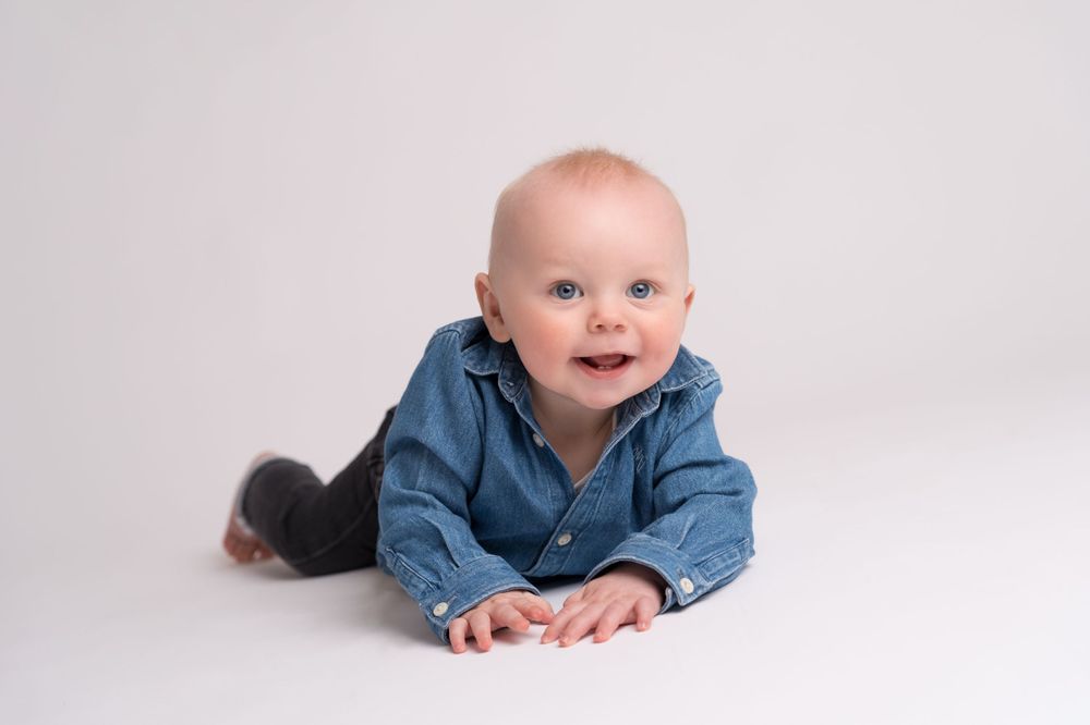 Baby wearing chambray shirt lies on white studio floor with happy expression.