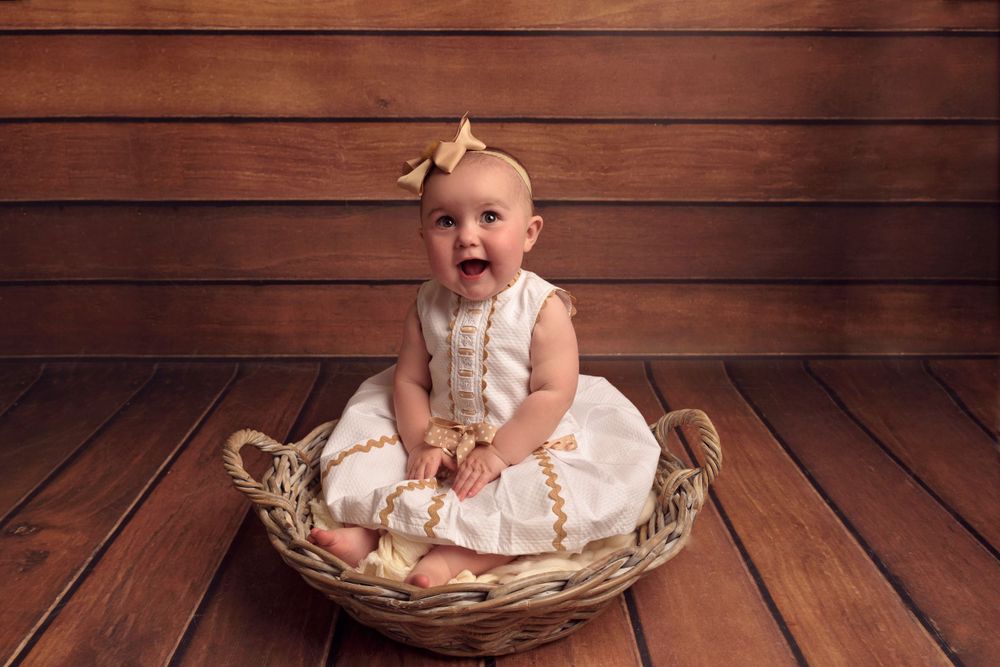 Small child in cream dress sits happily in wicker basket against wooden backdrop.