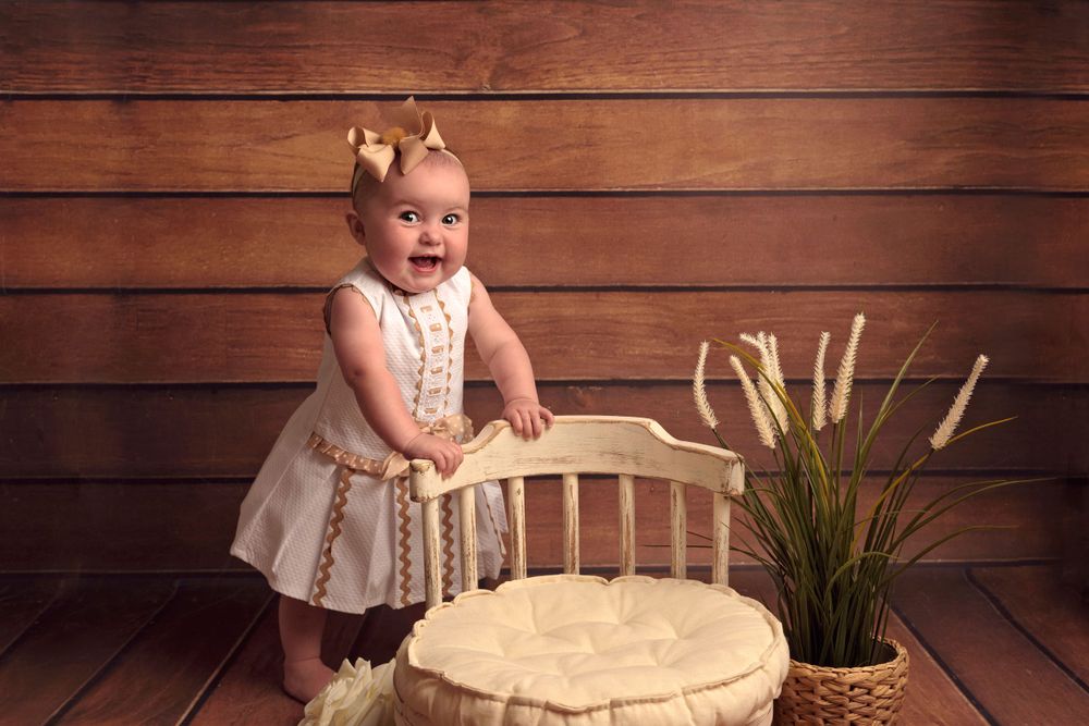 Small child in white dress stands beside rustic wooden chair and decor on brown wooden floor.