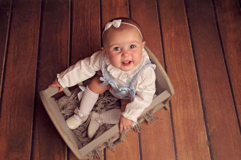 Small child in white outfit sits on vintage suitcase against wooden wall backdrop.