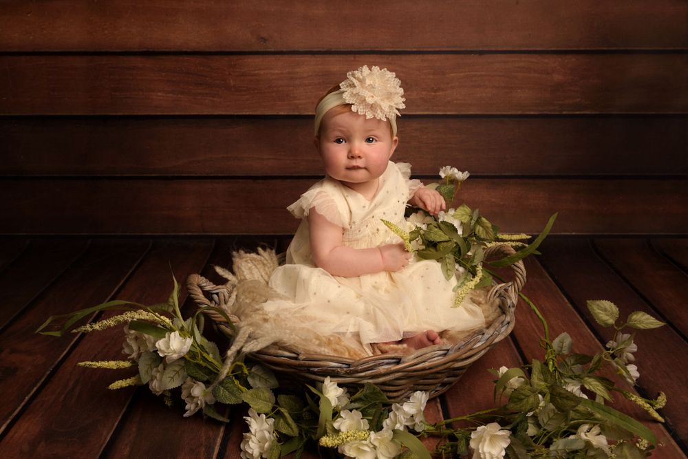 Small child in cream tulle dress sits in basket with floral decor against wooden backdrop.