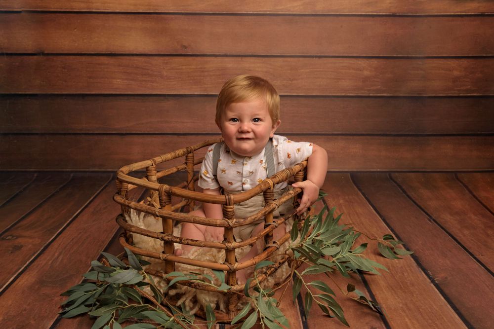 Child in white outfit sits in wooden crate with greenery against rustic backdrop.