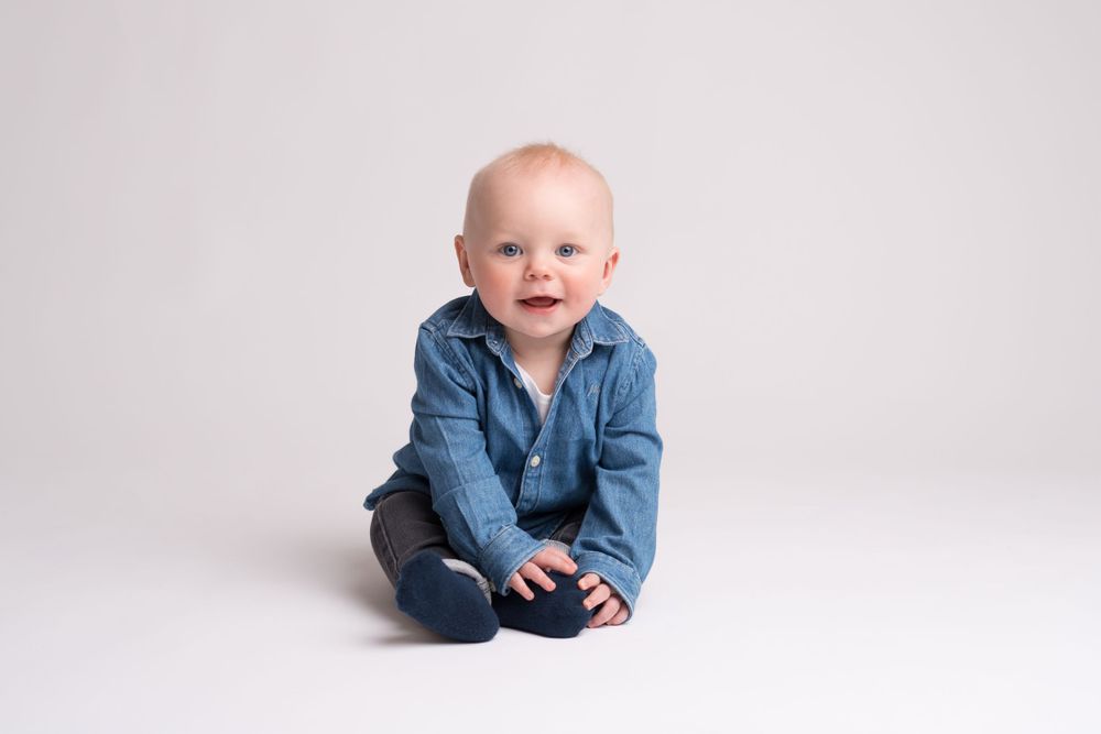 Baby in blue denim shirt sits on white seamless backdrop.