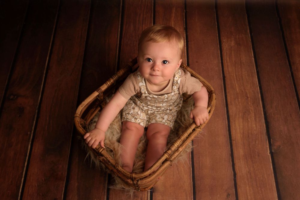 Small child in lace outfit sits in vintage wooden basket against dark wooden floor.