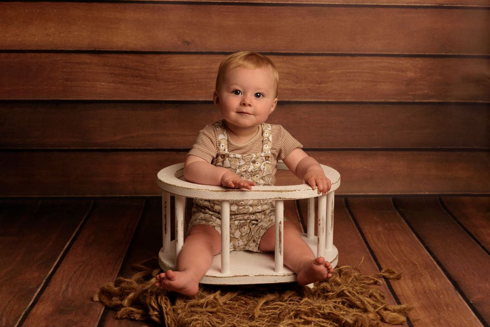 Baby in neutral-colored outfit sits in white vintage-style chair on wooden floor.