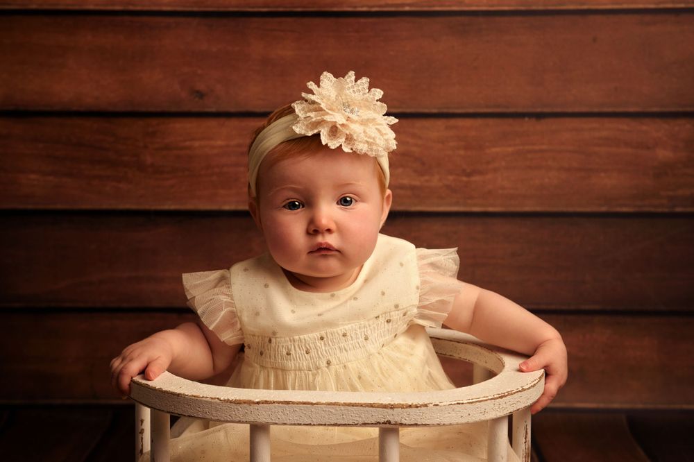 Small child in cream dress sits in vintage chair against wooden wall.