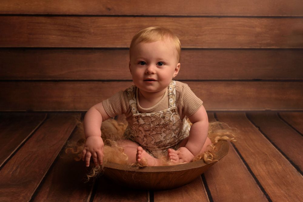 Small baby wearing beige overalls sits in a wooden bowl against rustic wooden wall backdrop.