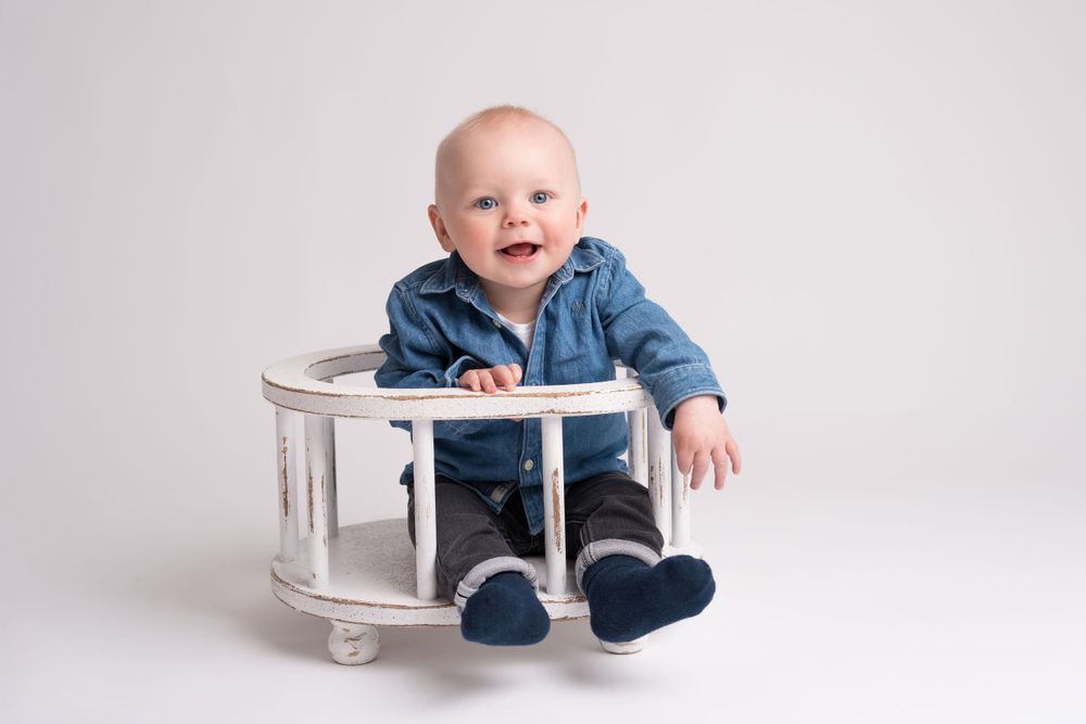 Baby wearing denim shirt sits in white round chair against light background.