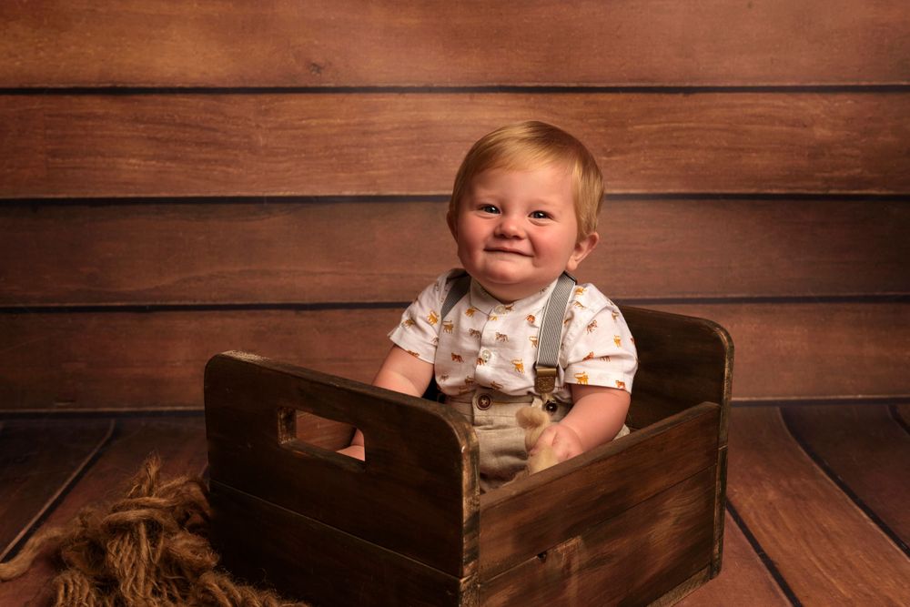 Smiling child in white outfit sits in rustic wooden crate against brown wall.