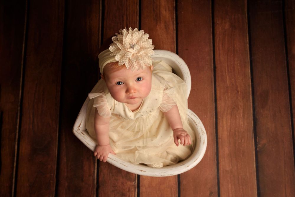 Small child in cream dress lies in white heart-shaped bowl against dark wooden backdrop.