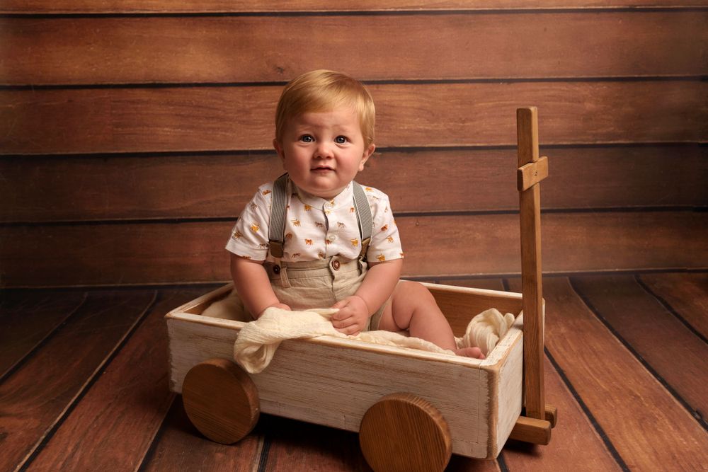 Smiling child in white outfit sits in wooden wagon against brown wall.
