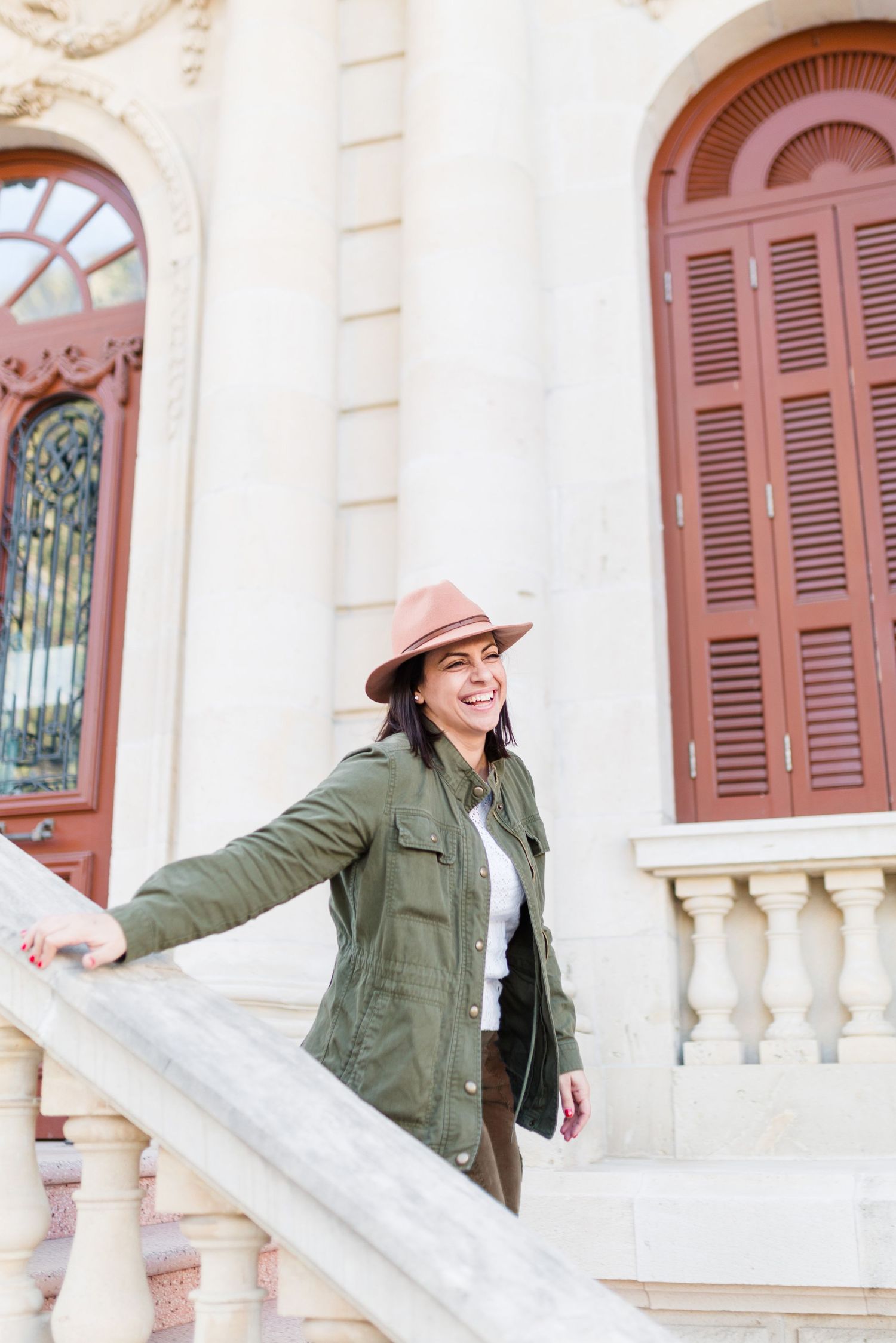 Woman in green jacket and hat stands on steps of building with ornate doors.
