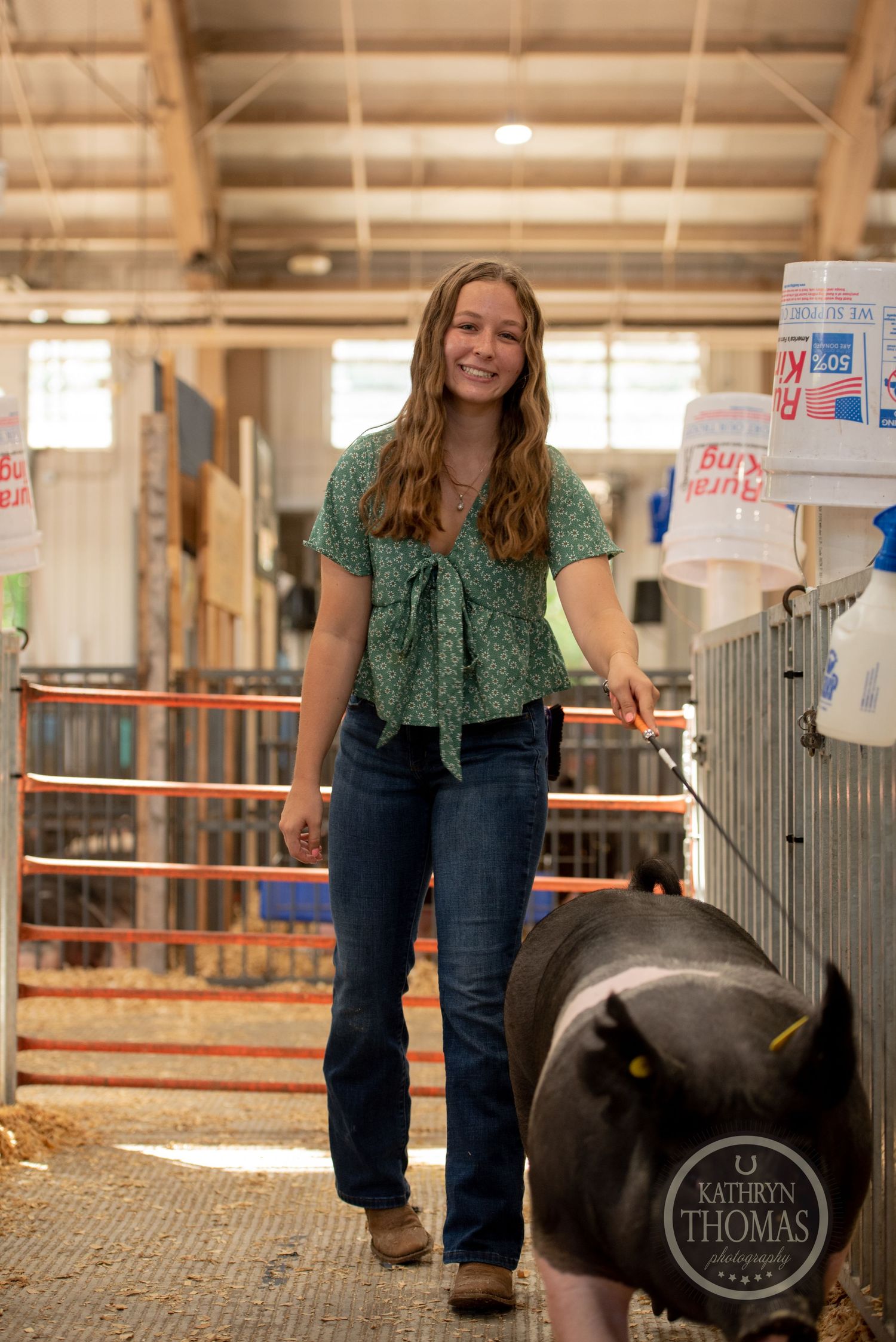 A person in green standing next to a black show pig in a barn walkway.