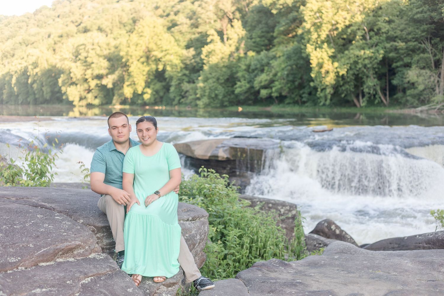 A couple poses together near a scenic waterfall in summer attire.