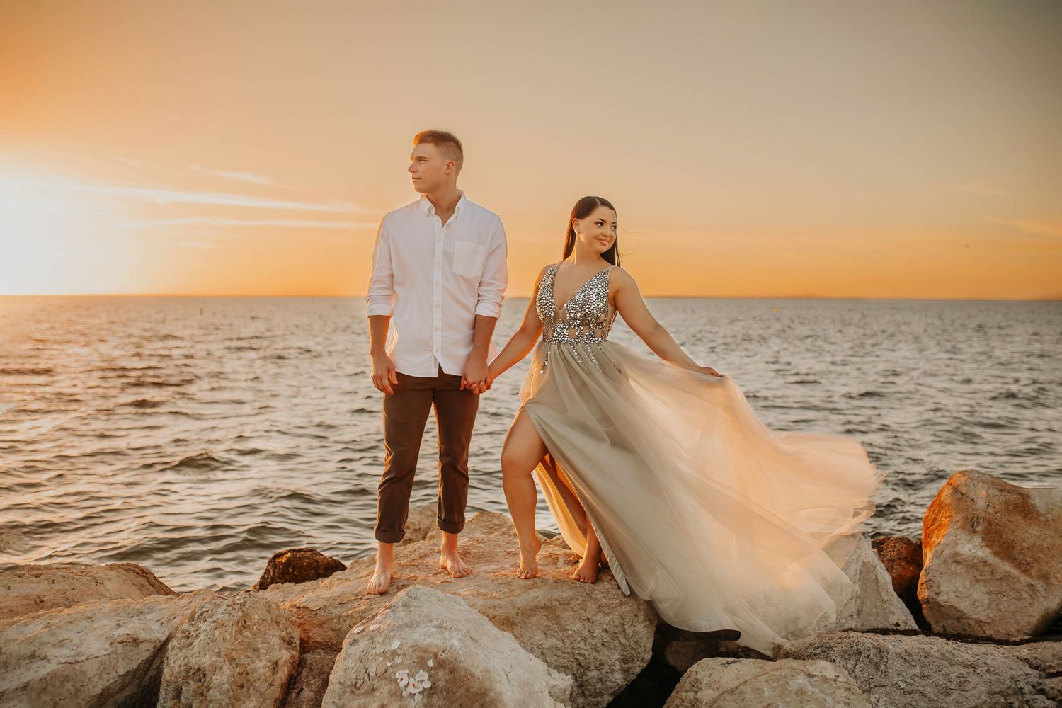 Couple holds hands on coastal rocks during golden hour as her elegant dress flows in the wind.