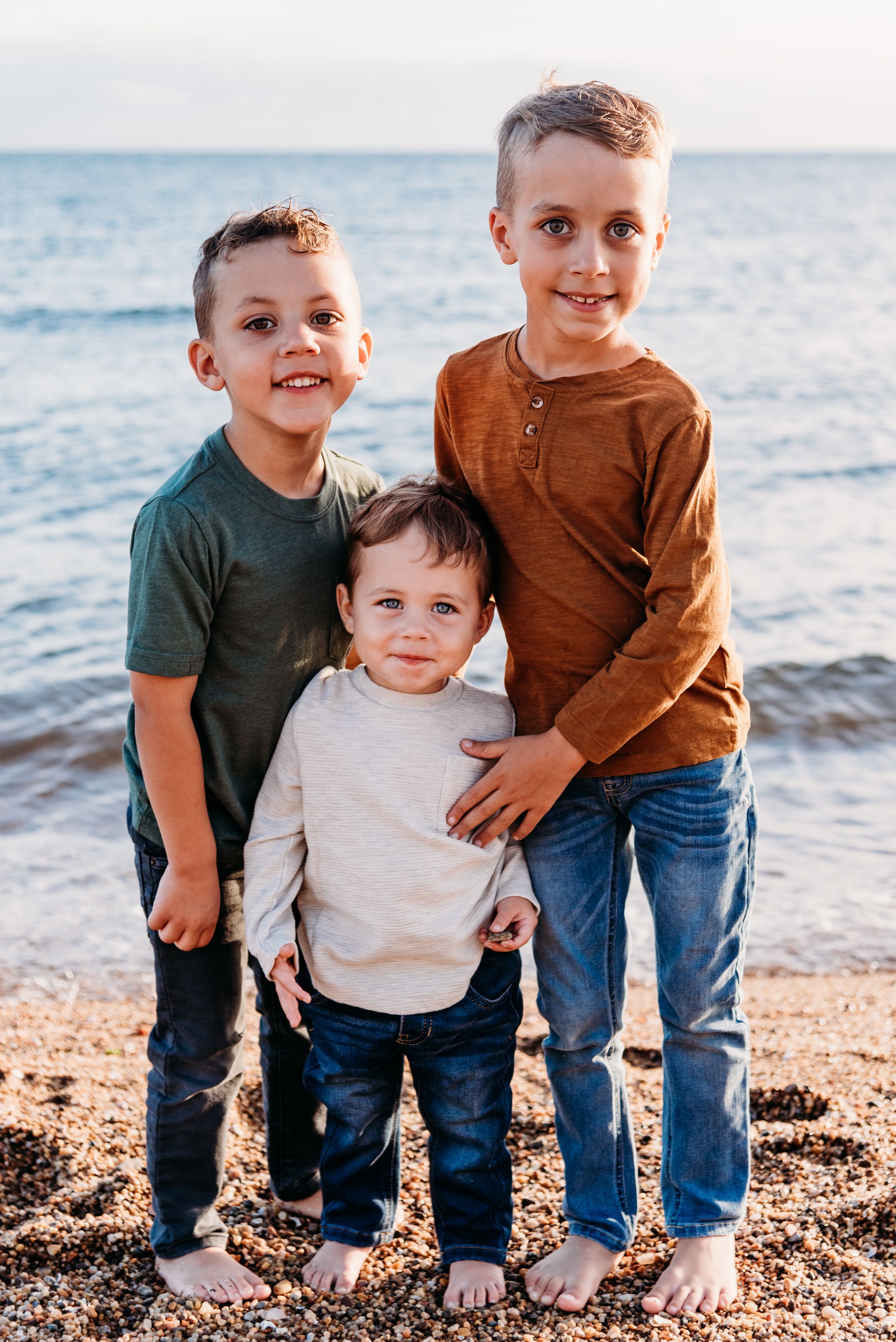 Three children standing together on beach at sunset wearing casual sweaters.