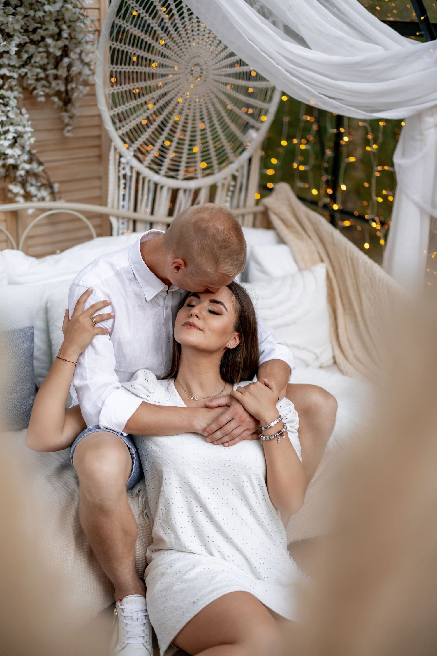 Romantic couple relaxing on white decorative furniture with fairy lights in background.