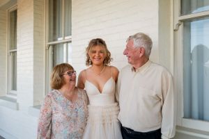 A bride in a white wedding gown poses with family members on a covered porch with white columns.