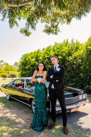 Couple in formal attire poses next to vintage black Cadillac under green trees.