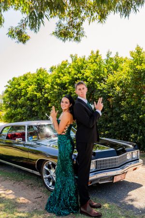 Playful formal couple strikes back-to-back pose by classic black car under trees.