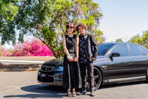 A dressed-up pair stands in front of a black luxury car with pink flowering bushes in the background.
