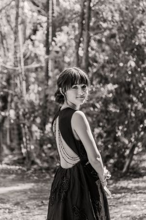 A black and white portrait of someone in an elegant backless dress posed in a wooded setting.