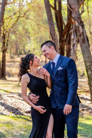 A couple in formal attire share an intimate moment in a sunlit forest setting.