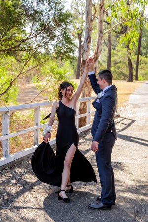A couple in elegant attire dance on a rustic path surrounded by trees.