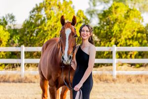 A beautiful chestnut horse stands with a woman in a black dress by a white fence.