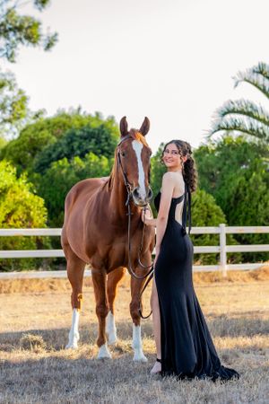 A woman in a flowing black dress poses with a horse against a summer landscape.