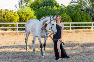 A woman in a black gown stands beside a dappled grey horse in a fenced paddock.