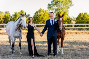 A couple in formal attire stands between two horses - one white and one brown - in a fenced field.