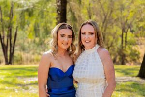 Two friends pose together outdoors in formal dresses against a sunlit nature backdrop.