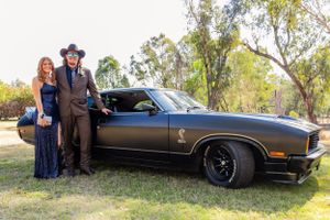 Couple in western attire poses beside classic black muscle car under trees.